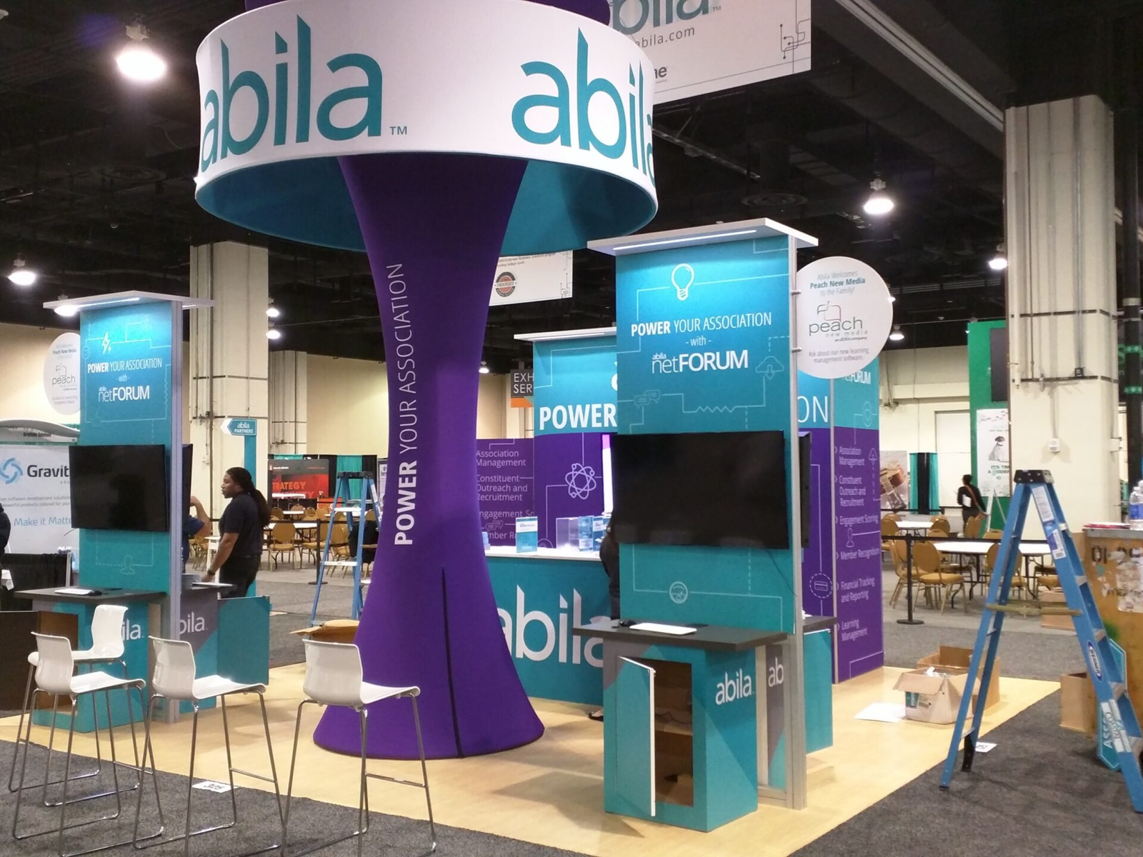 Abila 20x20 booth at a trade show under construction. Ladders and crates are in the photo. Large purple fabric funnel tower at the center of the display.