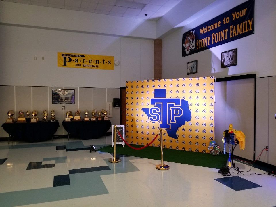 Photo Station in the Stony Point High School dining hall. A 10 foot backdrop with the STP logo centered on the display and a step and repeat background. This has some artificial turf in front of it and trophies in the background. A great spot for families to get photos in front of this display.