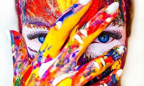 Artistic closeup photo of a person with their hand over their face. The entire person is covered with splattered multi-colored paint.