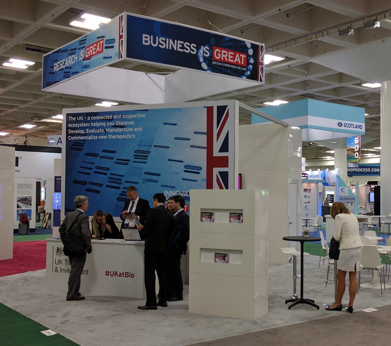 The United Kingdom Trade and Industry booth at a trade show. Large wall with a reception counter in front of it. There are a few people in business suits having conversations. A 10ft hanging box sign is over the booth that reads "Business is Great".