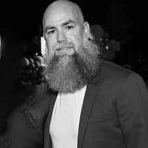 Black and white photo of Travis Wilky. He is bald and have a large expansive beard. He is wearing a light colored tshirt and dark sport coat.