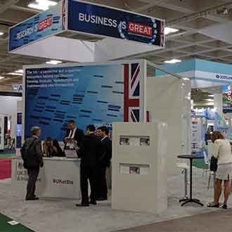 The United Kingdom Trade and Industry booth at a trade show. Large wall with a reception counter in front of it. There are a few people in business suits having conversations. A 10ft hanging box sign is over the booth that reads "Business is Great".