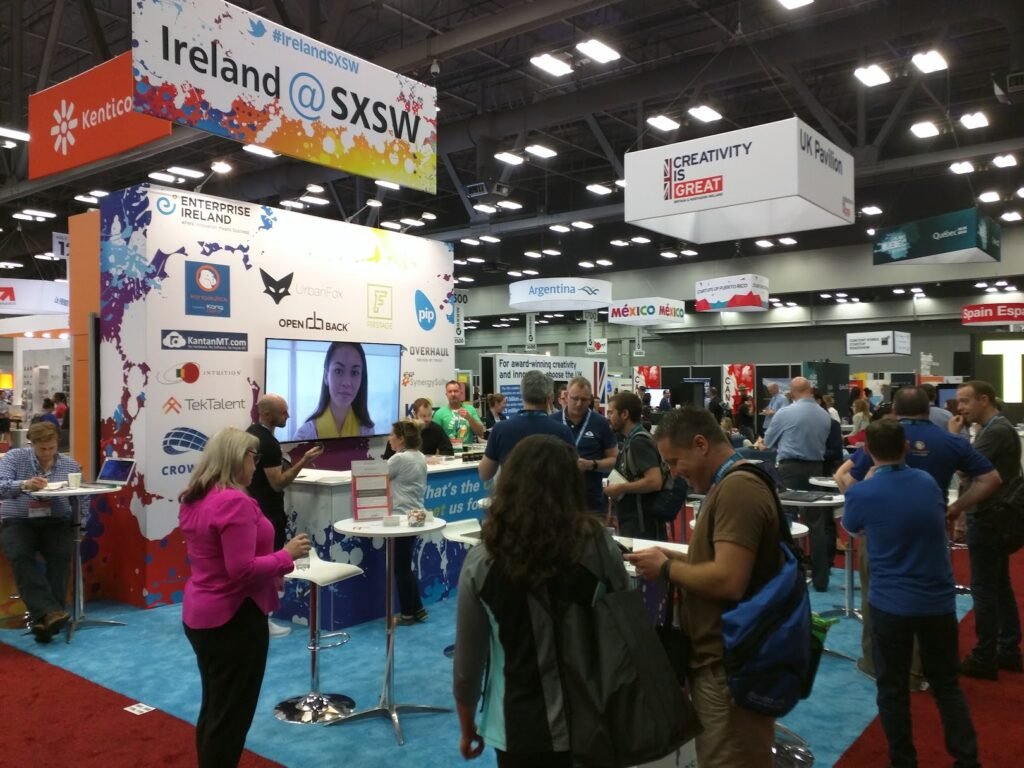 Ireland @ SXSW 20x20 trade show booth. Blue carpet with many people having conversation. Large wall with television monitor near the back of the booth and a flat hanging sign with colorful imagery and reads Ireland @ SXSW.